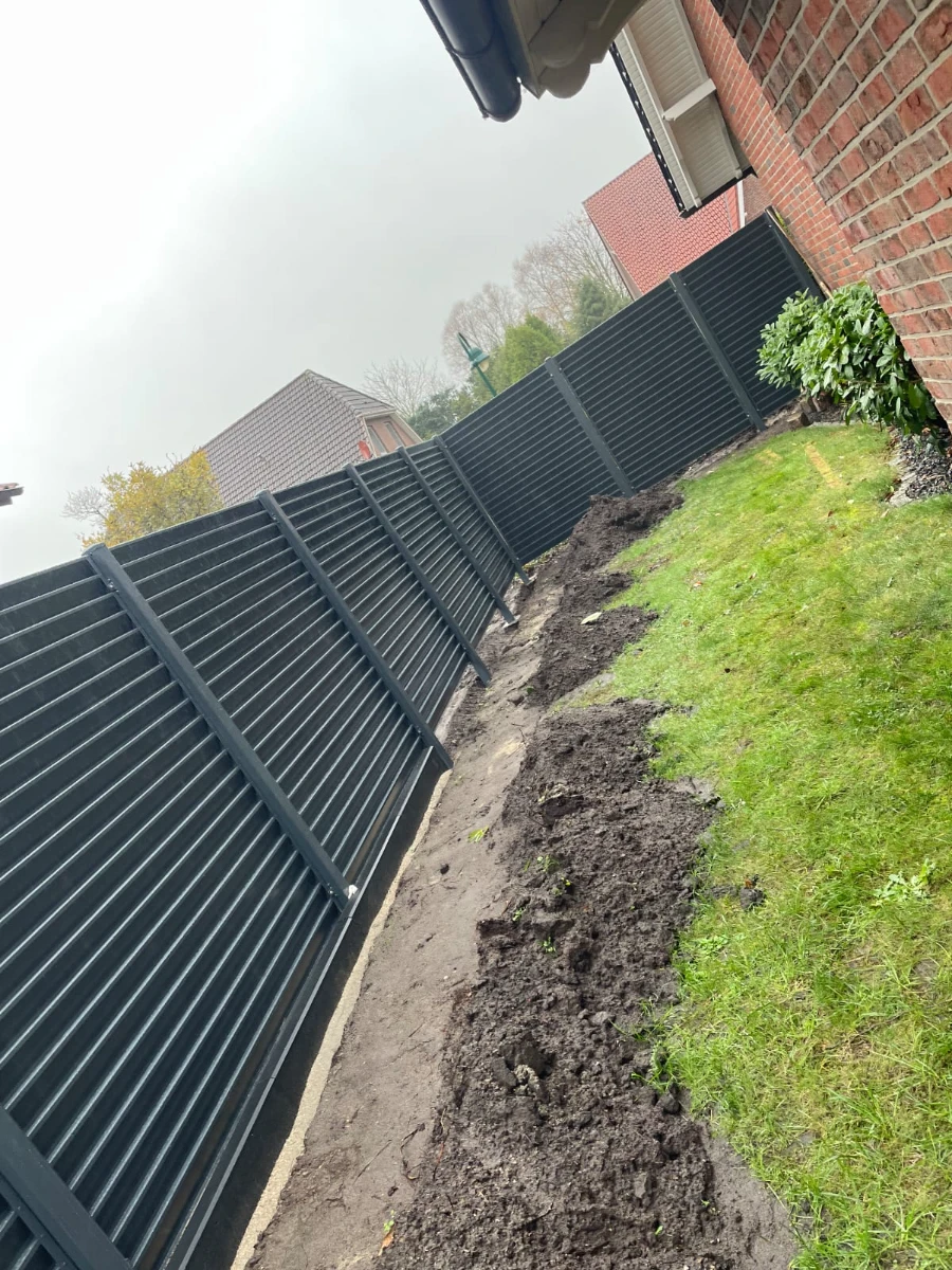 Dunkler Aluminium-Lamellenzaun entlang Hauswand mit Rasen und frisch aufgeschütteter Erde