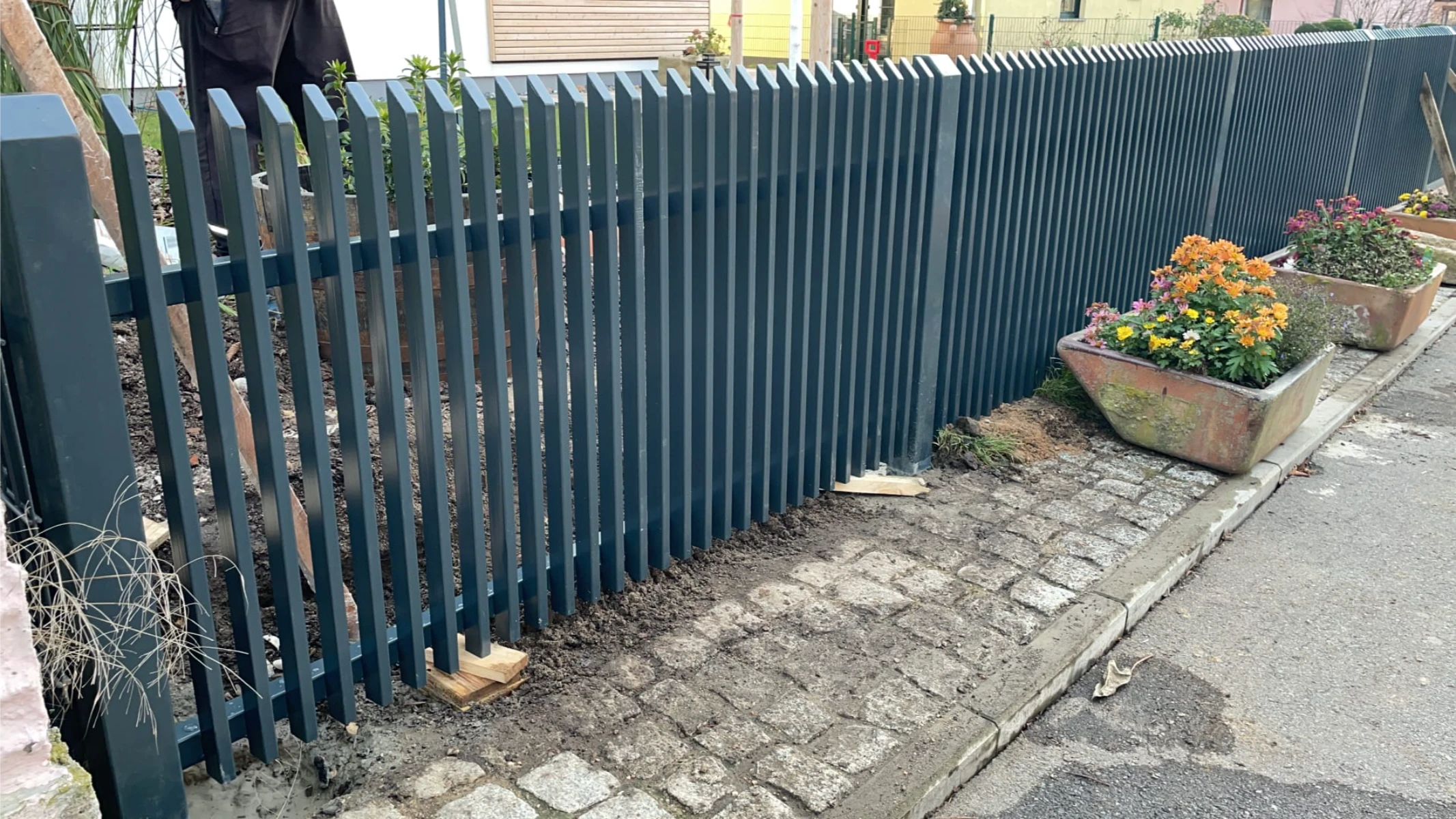 Dunkler Lamellenzaun aus Metall entlang Pflasterweg, daneben bepflanzte Blumentröge