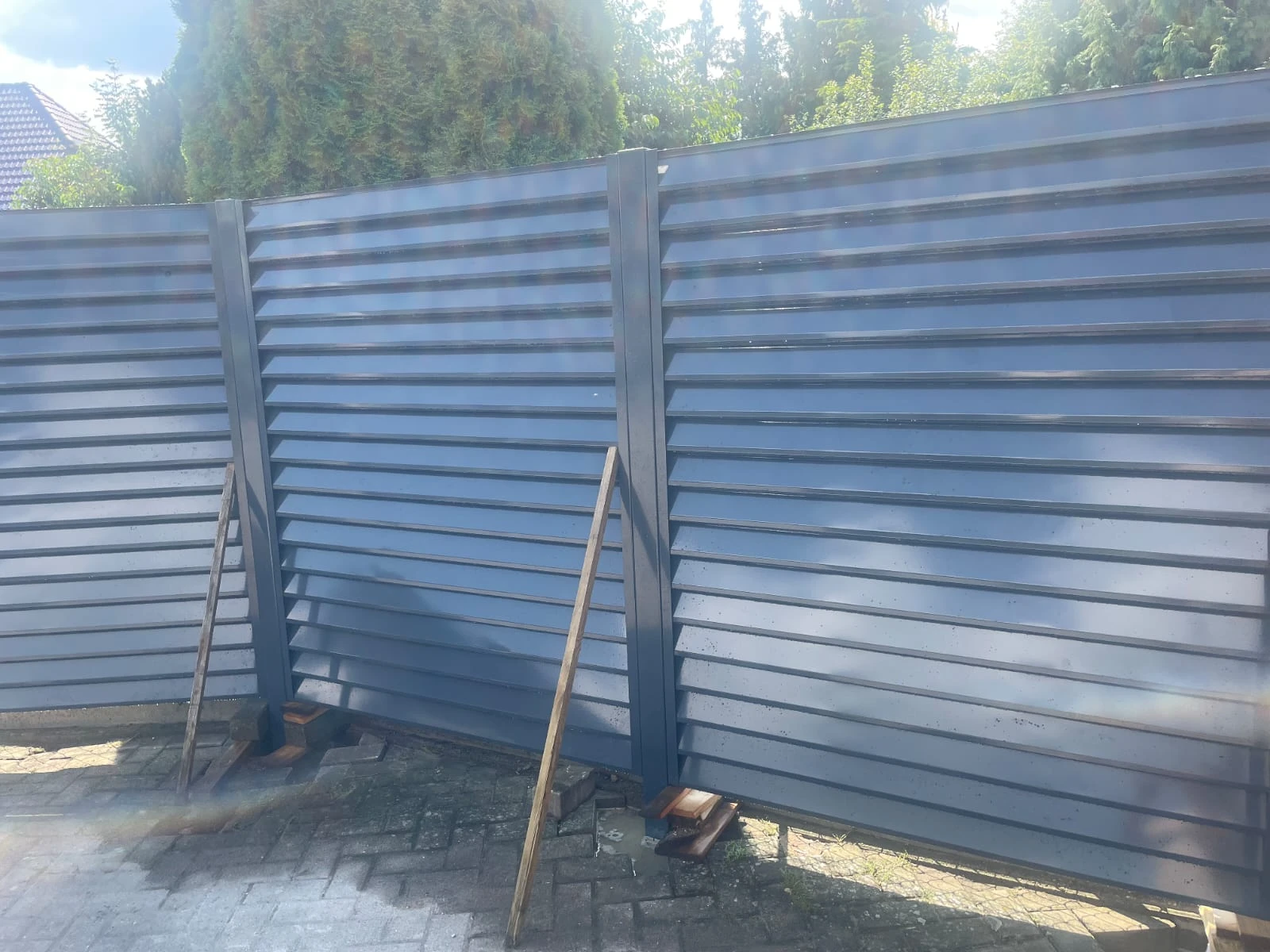 Provisorisch abgestützter Lamellenzaun aus pulverbeschichtetem Aluminium vor gepflasterter Einfahrt