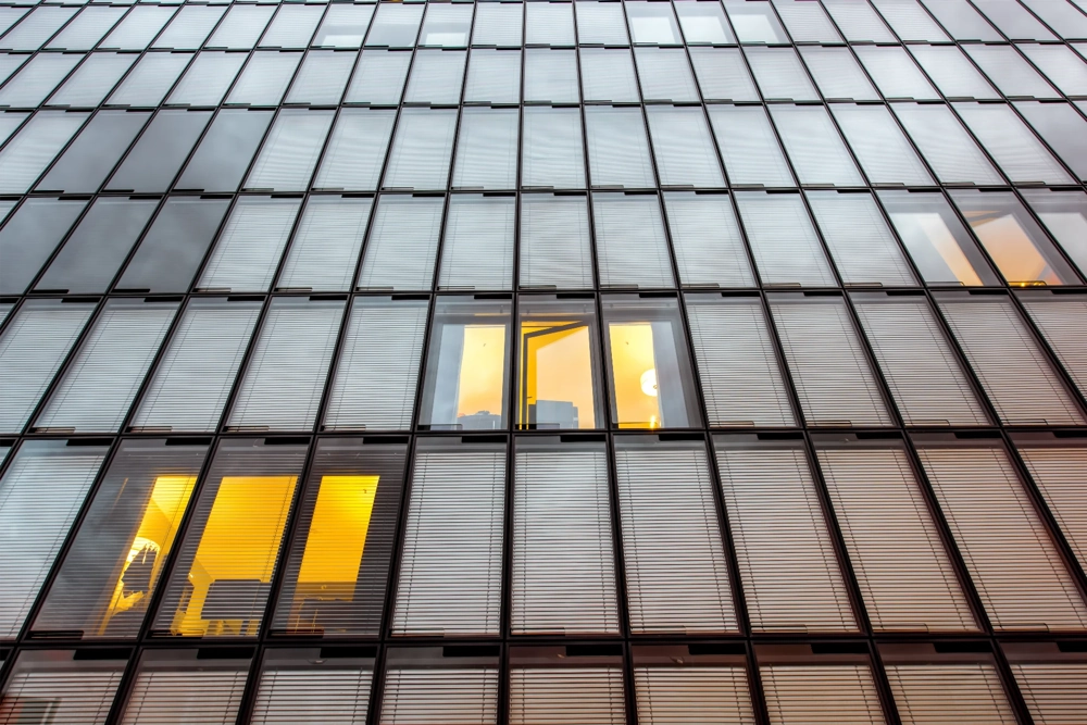 Moderne Glasfassade mit regelmäßigen Fensterelementen und schlichten Außenjalousien, ein geöffnetes Fenster strahlt warmes Licht aus